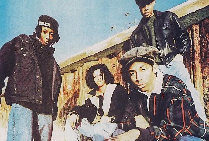 Four artists in casual streetwear pose outdoors before wooden structure under clear sky with relaxed energy.