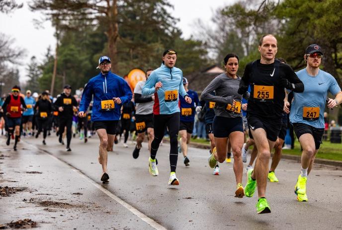 Runners in a road race move along a wet street, wearing numbered bibs and running outfit.