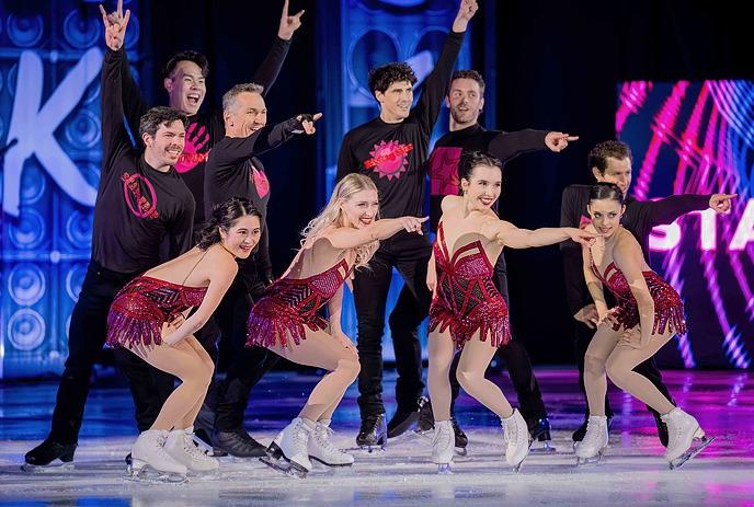 Ten skaters pose on ice in red and black costumes, smiling under blue and purple lights.