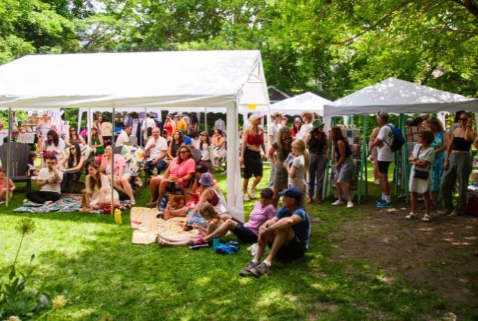 Shaded park festival with tents, people relaxing on grass, and crowds wandering under tall green trees.