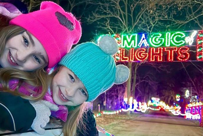 Two children in colorful winter hats pose near a glowing Magic Lights display at night.