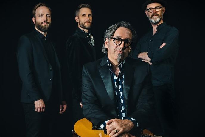 Four artists dressed formally pose with a guitar before a sleek black backdrop patterned.