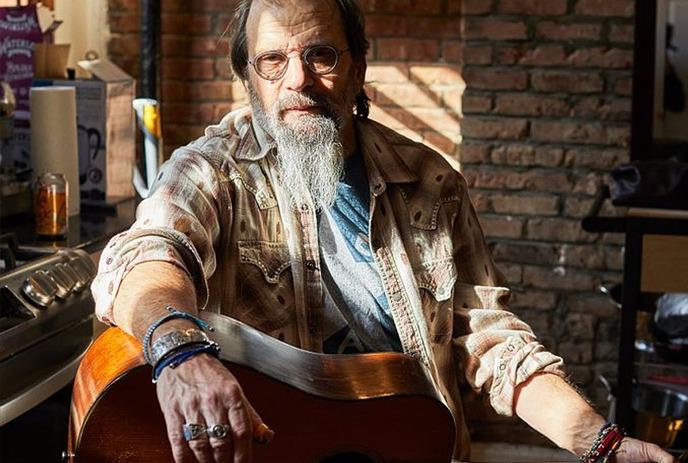 Steve Earle wearing a patterned shirt and denim jacket, adorned with multiple rings, is holding an acoustic guitar.
