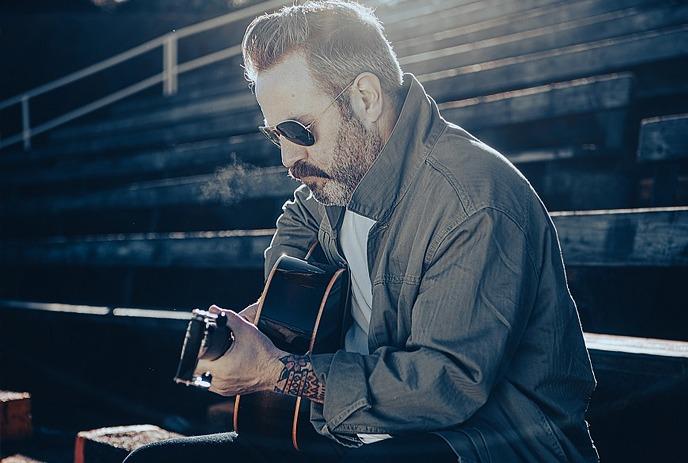 Artist in sunglasses plays acoustic guitar on outdoor under a dramatic sunlight, tattoos, cold breath visible.