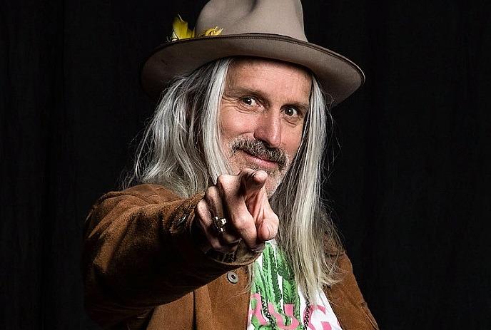Steve Poltz wearing a hat, smiling, and pointing his right hand at the camera, in front of a black background.