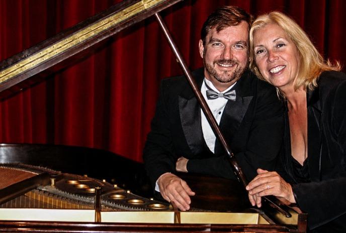 Clark Bryan & Marion Miller poses beside a grand piano in formal attire, with elegant red curtains on the background.