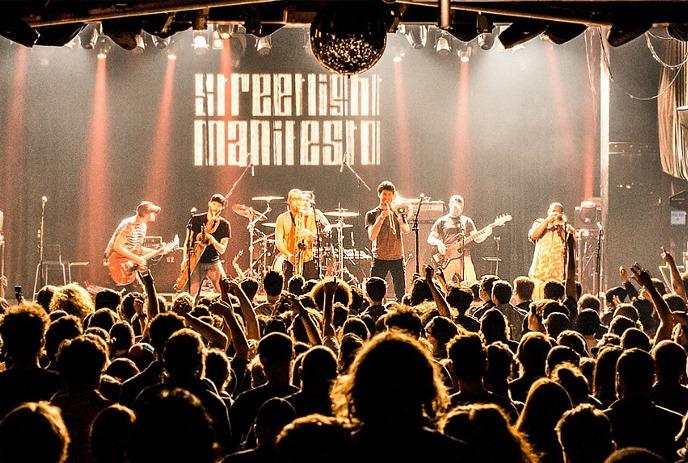 A packed crowd faces a band onstage, bathed in warm spotlights as hands rise and music fills the venue.
