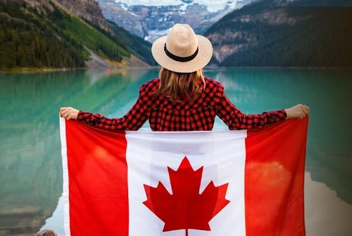 A woman wearing a plaid shirt and hat holds a Canadian flag facing a serene lake surrounded by mountains.