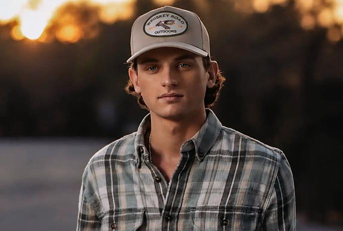 Person in plaid shirt and cap standing outdoors near a blue structure at sunset, hands in pockets.