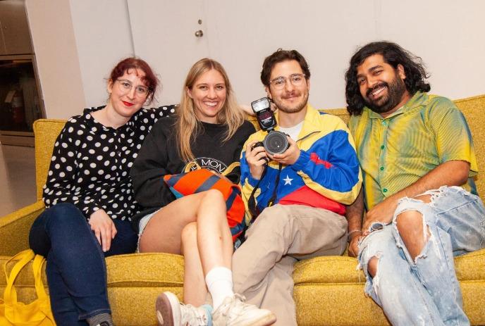 Four friends in eclectic outfits relax on a yellow couch, one holding a camera flash.