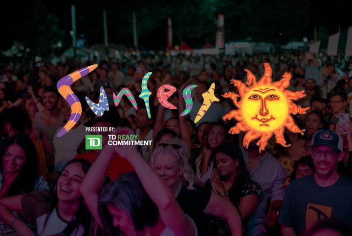 People watch a live band perform on a stage in a park under trees, with a colorful “Sunfest” backdrop and banner.