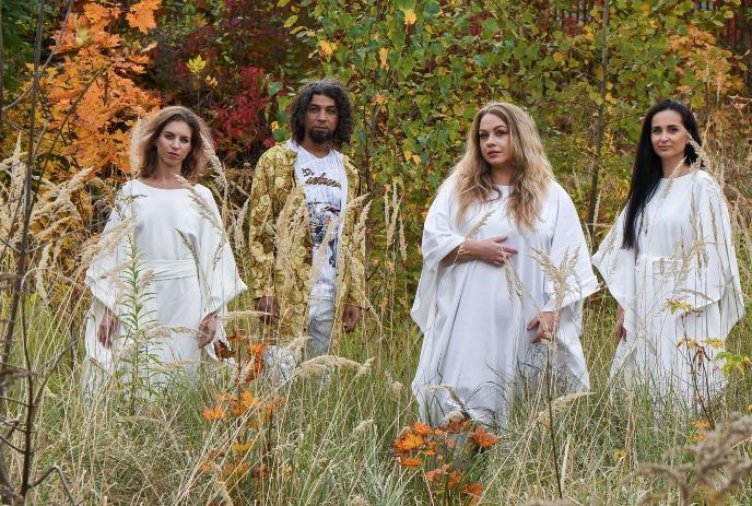 A group of four people in white outfits stand outdoors in tall grass with colorful autumn trees behind them.