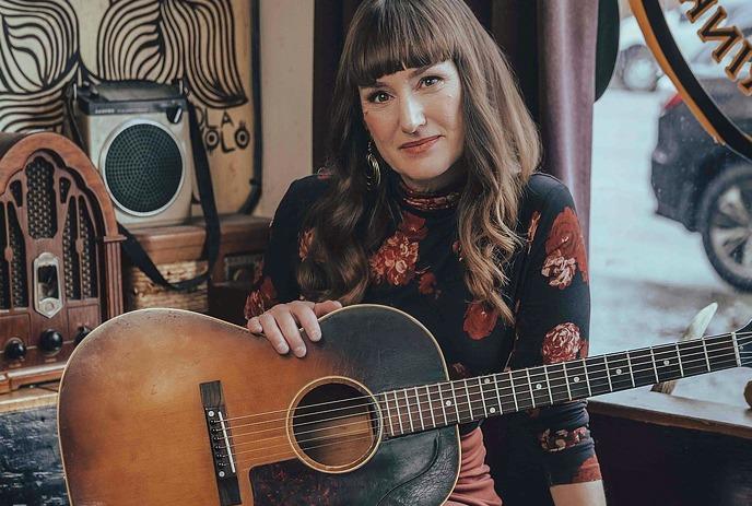 Suzie Ungerleider sitting down in a room, holding a guitar and looking at the camera.