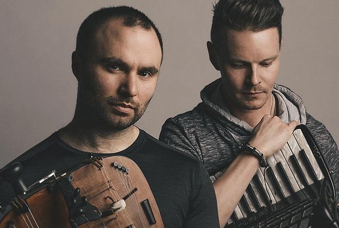 Two musicians pose in a studio with instruments against a plain backdrop.