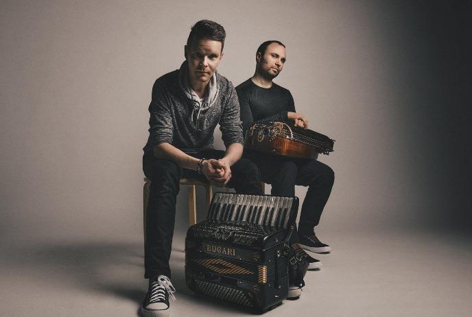 Two musicians pose in a studio with instruments against a plain backdrop.