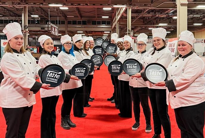 A group of chefs in white uniforms joyfully dancing on a red carpet at a bustling food event.