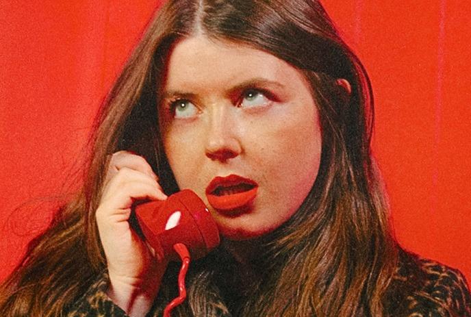 Lady artist in a red top holds a white electric guitar; background includes a guitar case and coffee cup.