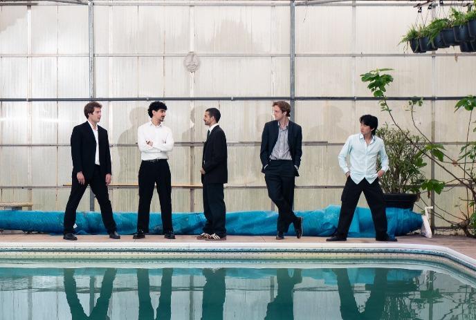 Five artists in formal wear pose by an indoor pool with plants and a rolled-up blue pool cover behind them.