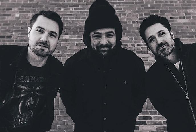 Three artists leaning forward in black and white portrait standing before brick wall in close formation.
