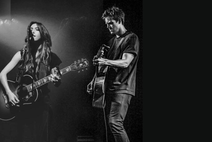 A black and white photo of Tyler Hilton & Kate Voegele on stage playing their guitars and singing.