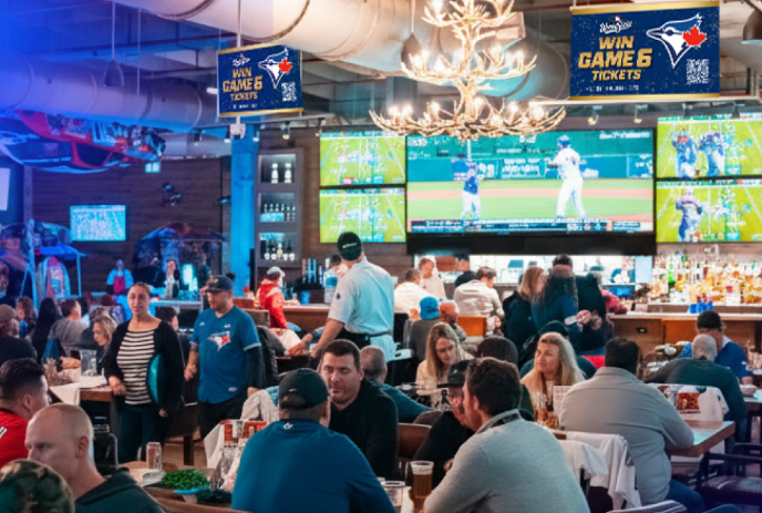 Fans in Blue Jays gear watch baseball on big screens in lively sports bar with rustic decor and promos.