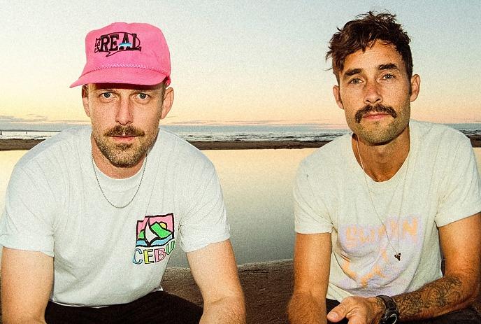 Artists kneeled on the beach at sunset wearing casual shirts and caps beside calm water in warm light