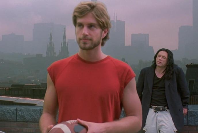 A man holding a football on a rooftop with a city skyline in the back, with another man behind him