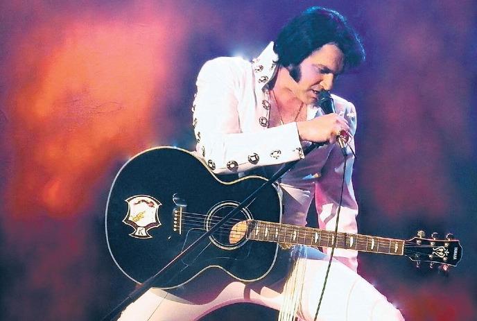 Tim 'E' Hendry in costume as Elvis, performing with a microphone and guitar on stage.
