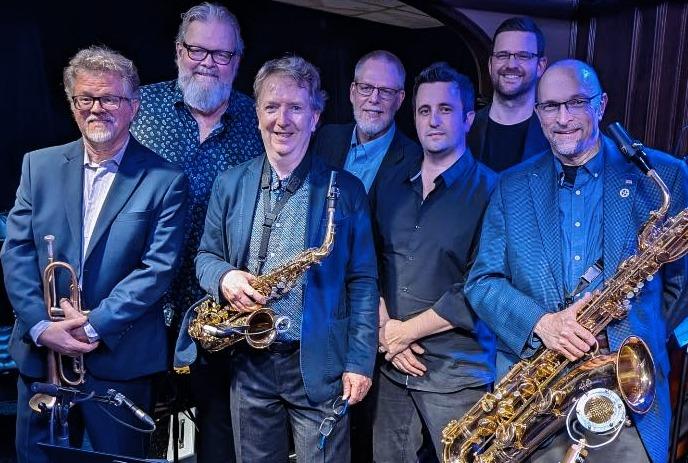 A group of seven men dressed in suits, smiling on stage with musical instruments.