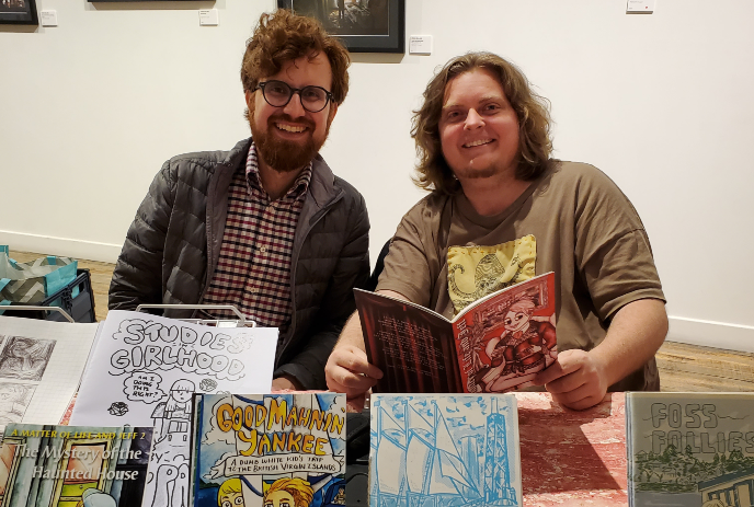 Two artists sit at a table displaying illustrated books and comics with framed art behind them.