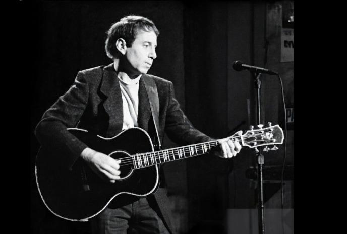 A black and white photo of Paul Simon playing the guitar on stage with a microphone in front of him.