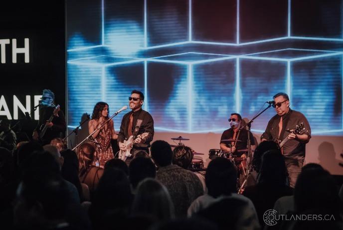 Crowd watches as a band on lit stage as musicians perform before a bright blue geometric screen backdrop.