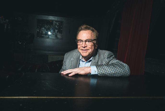 Tom Arnold with glasses and a checkered blazer smiles warmly in a dimly lit room, leaning on a black surface.