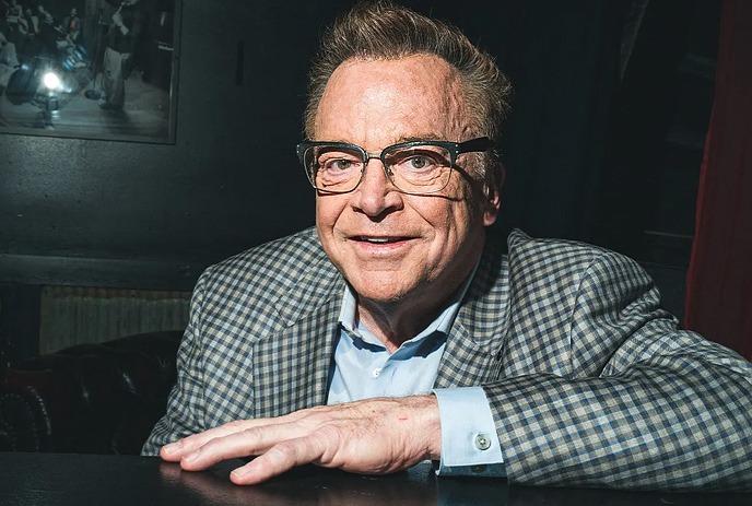 Tom Arnold with glasses and a checkered blazer smiles warmly in a dimly lit room, leaning on a black surface.