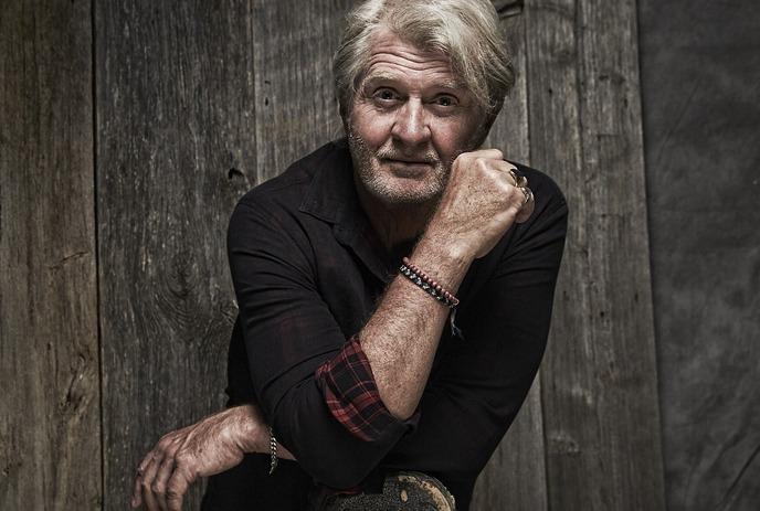Person with light hair in dark shirt with plaid cuffs, leaning on wood surface, rustic wooden backdrop behind.