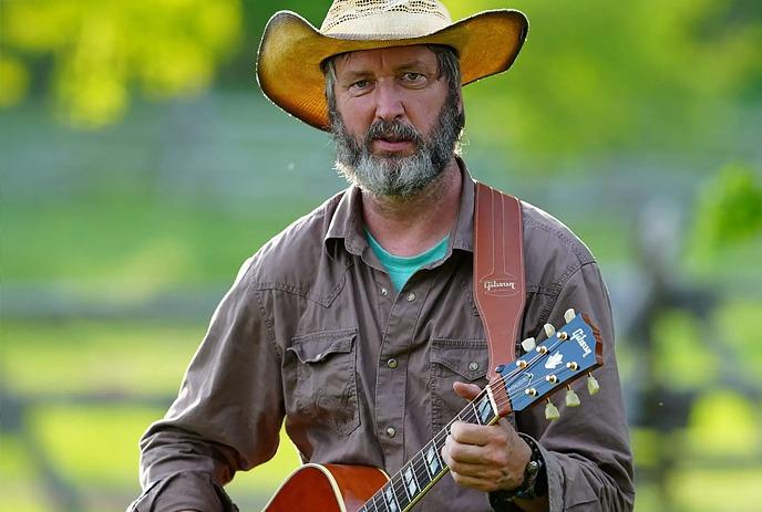 Tom Green wearing a cowboy hat and playing guitar, in a field on a sunny day.