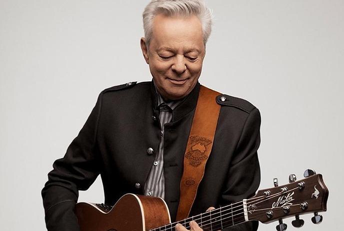 Tommy Emmanuel wearing a suit while playing his guitar against a white background.