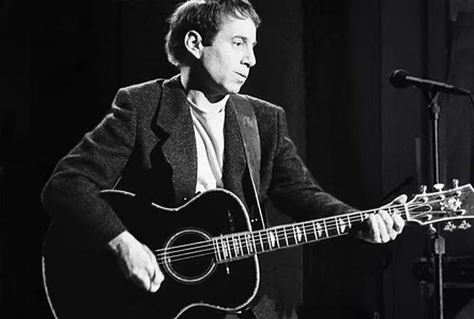 A black and white photo of Paul Simon playing the guitar on stage with a microphone in front of him.