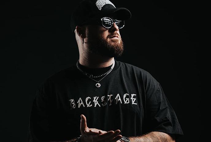 Artist in black backstage tee and cap stands against a dazzling starry night sky backdrop.