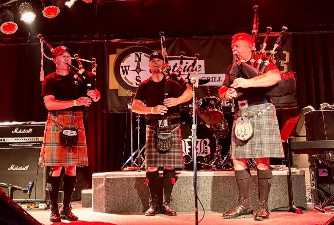 Three kilted players perform bagpipes on red?lit stage with drum set and amps behind them.