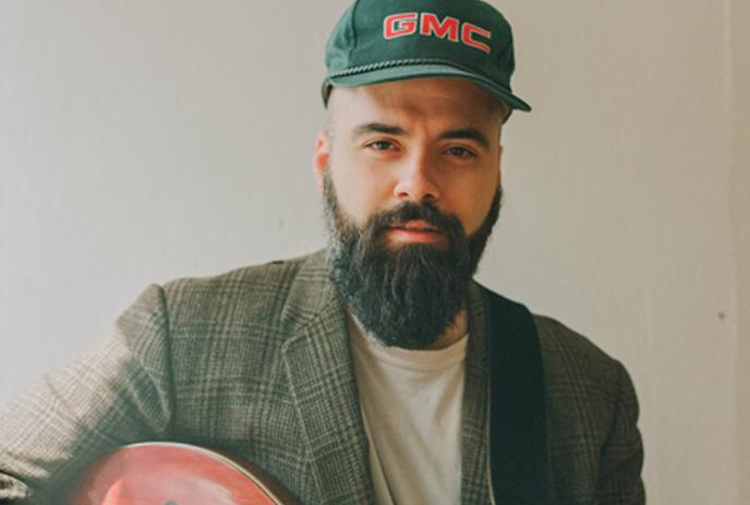 Musician in plaid jacket and green cap holds guitar while posing against white background.