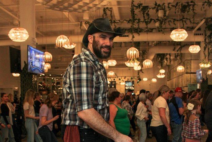 A man wearing a cowboy hat dances in a lively room filled with people, adorned with hanging lights.