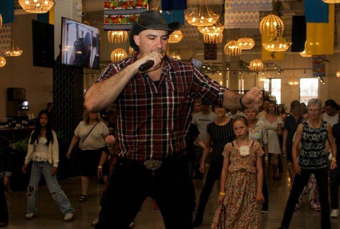 Urban Cowboy line dancing with a crowd in the background, wearing a cowboy hat and flannel shirt.