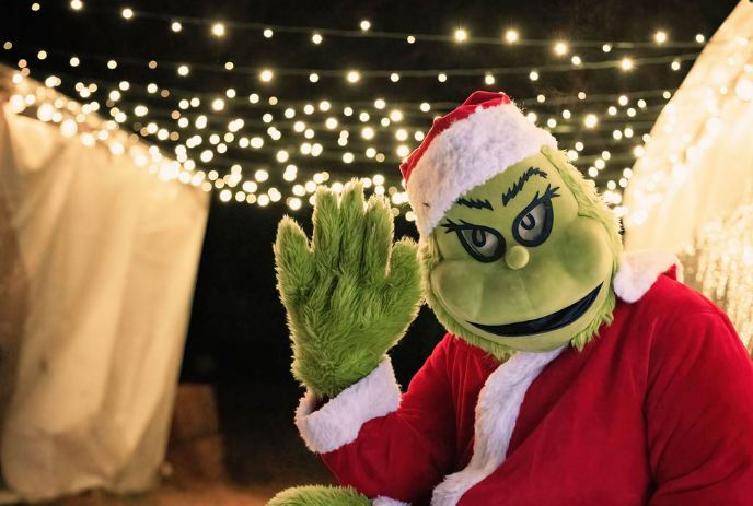 Grinch in Santa outfit waves cheerfully under glowing string lights at a festive nighttime holiday event.
