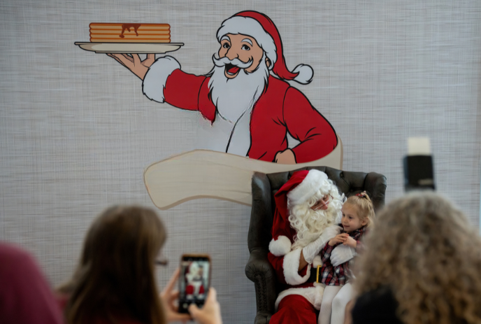 Santa holds child in chair as people take photos with a pancake Santa mural in the background.
