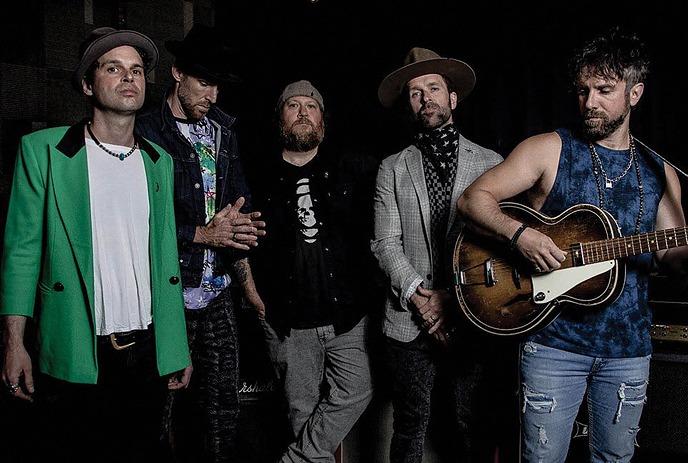 Five musicians pose in dim studio, one playing acoustic guitar, posing for the picture.