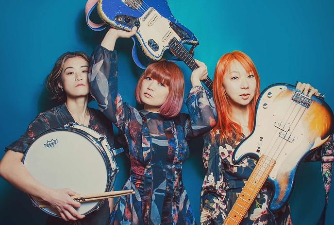 Three women artists with two guitars and one drum posing for a picture under a blue background.