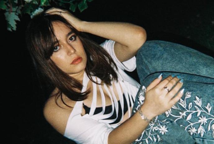 Artist Lelê seated outdoors wearing a cutout white top and embroidered jeans.