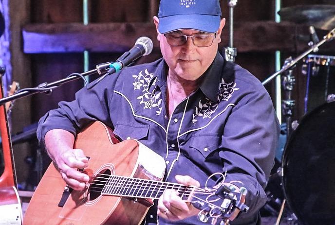 Musician Pete Denomme in glasses and a cap beside a microphone stand.
