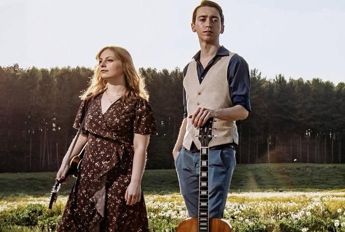 Two musicians with guitars standing in a flower-filled field beside an open guitar case.